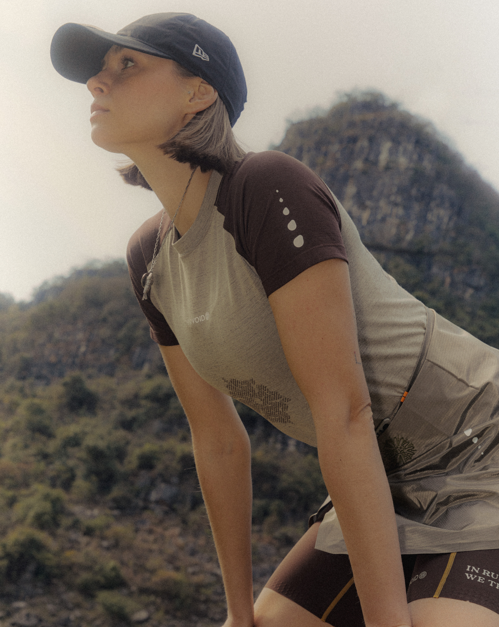 A person wearing a UPPERVOID cap and form athletic wear leans forward outdoors, with green hills and
rocky mountains blurred in the background under a bright sky.