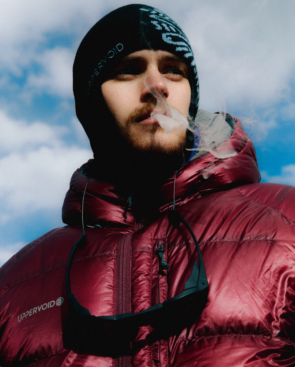A person wearing a UPPERVOID cap and form athletic wear leans forward outdoors, with green hills and 
            rocky mountains blurred in the background under a bright sky.