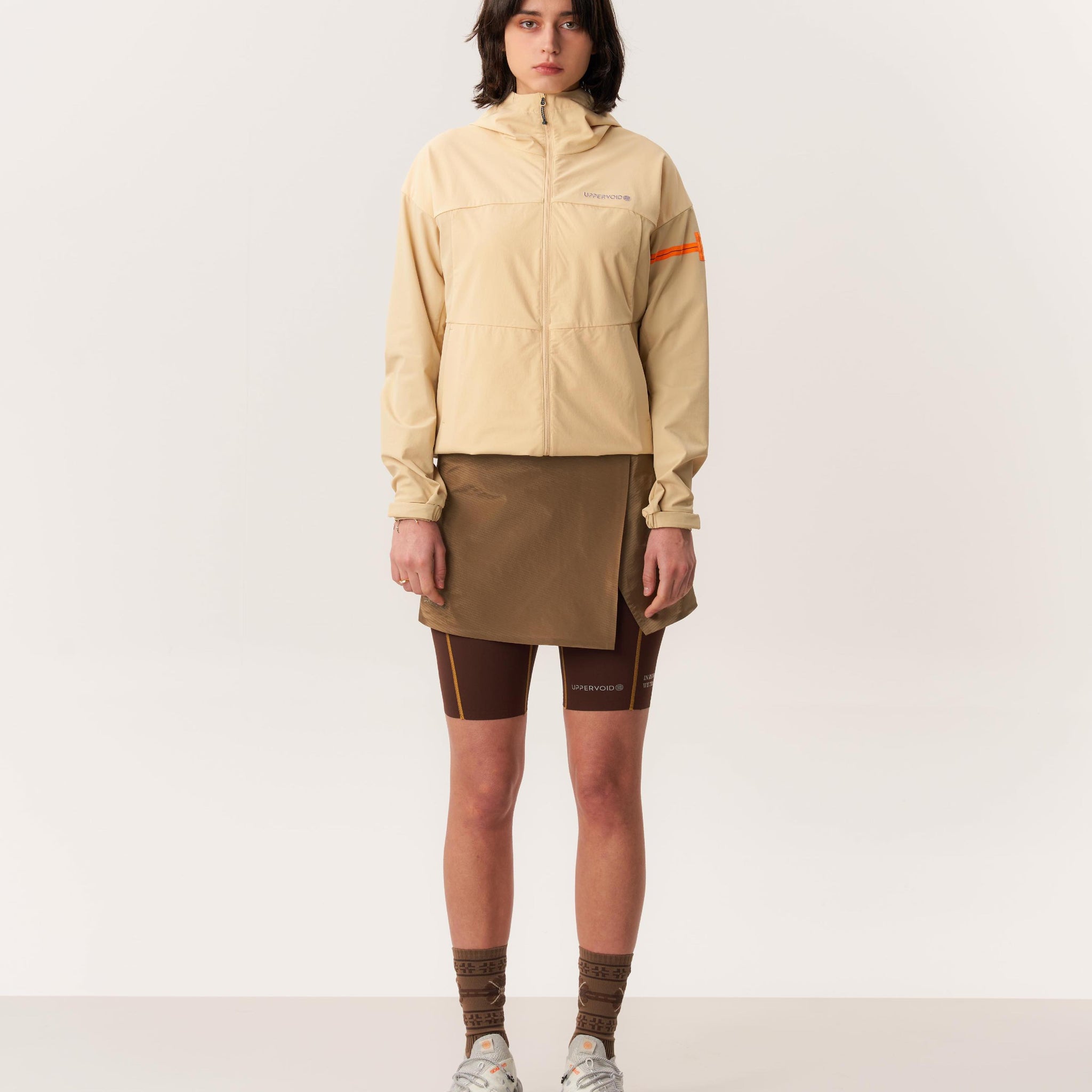 Model wearing beige packable rain jacket for hiking with light brown hardshell skirt in studio lighting