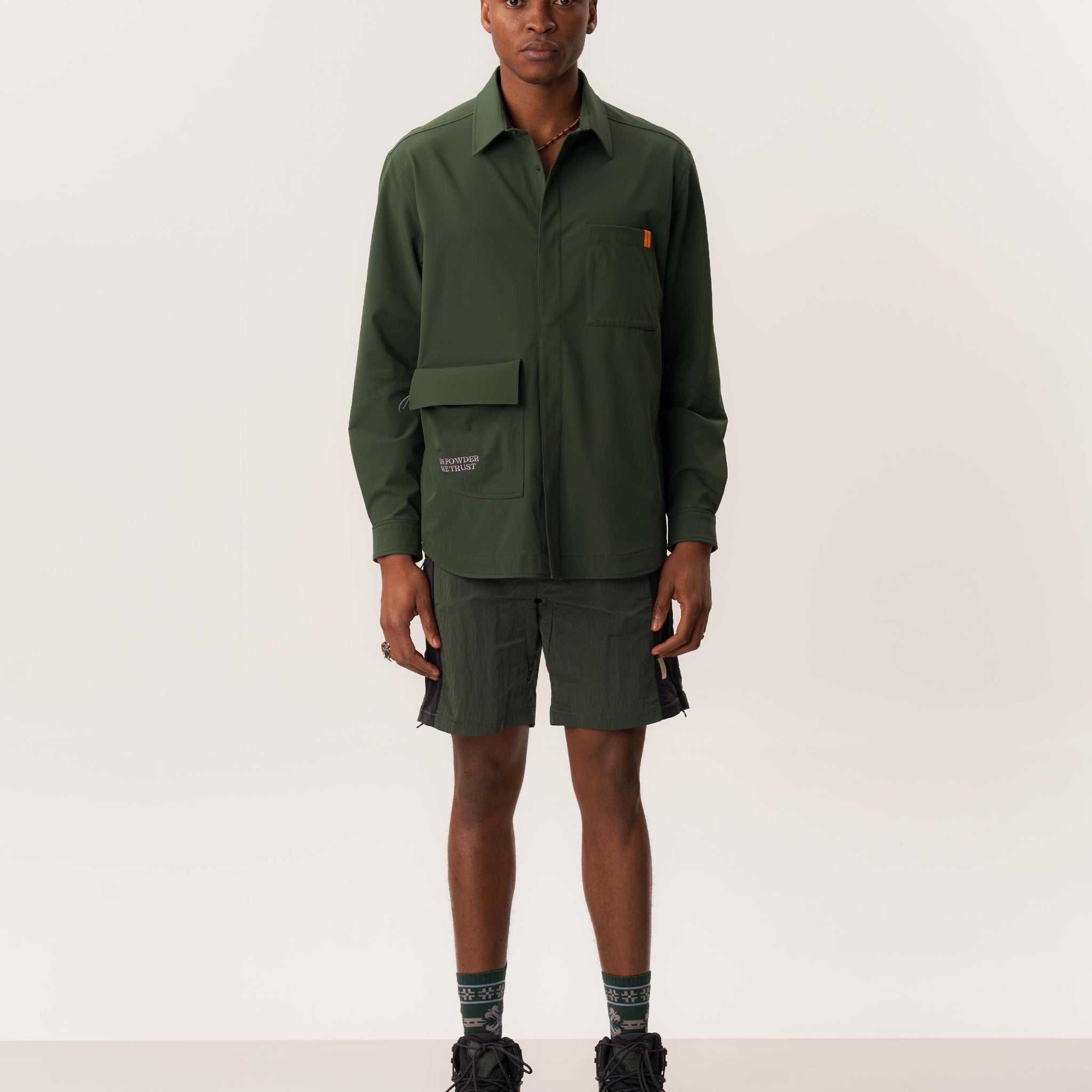 Model wearing dark green overshirt and green wrinkled shorts in clean studio environment