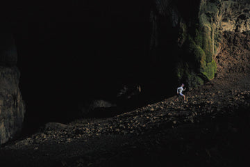 Model in neutral-toned hiking gear standing near the mouth of a moss-covered cave