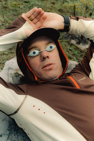 Model resting on rocky surface, wearing a hoodie and playful goggle-style eyewear