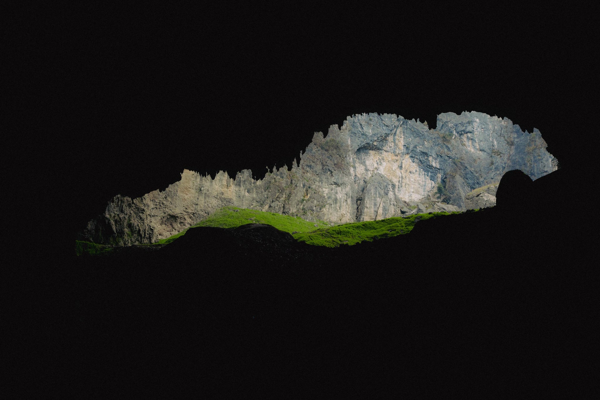 Cave framing rugged stone mountains and vibrant grasslands under midsummer light